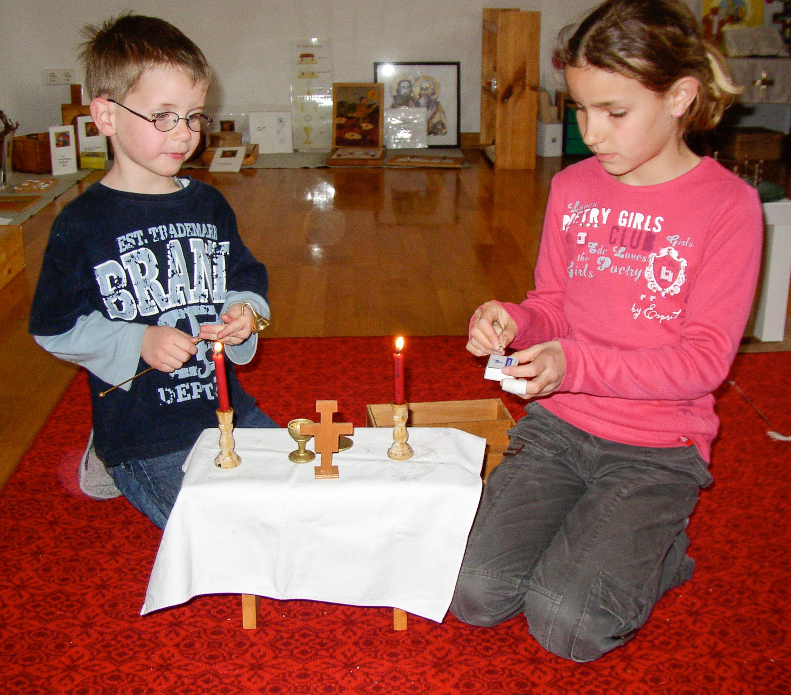Zwei Kinder haben das Modell vom Altar mit Hostienschale, Kelch, Kreuz und Kerzen aufgebaut, ein Kind zündet die Kerzen an.