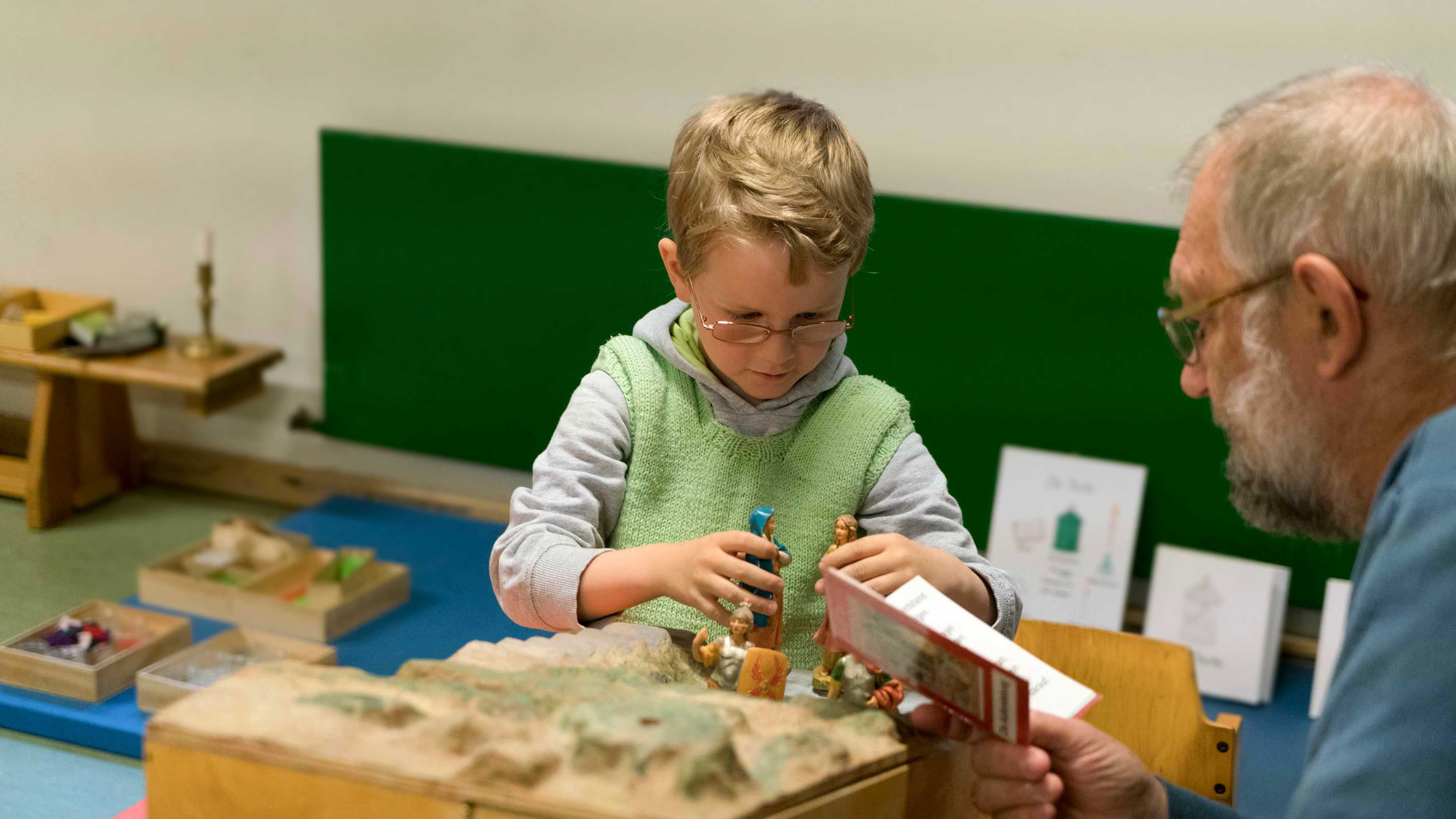 Ein kleines Kind arbeitet gesammelt mit den Figuren der Frauen, die entdecken, dass das Grab leer und Jesus auferstanden ist. Ein Mann liest ihm den Bibeltext vor.