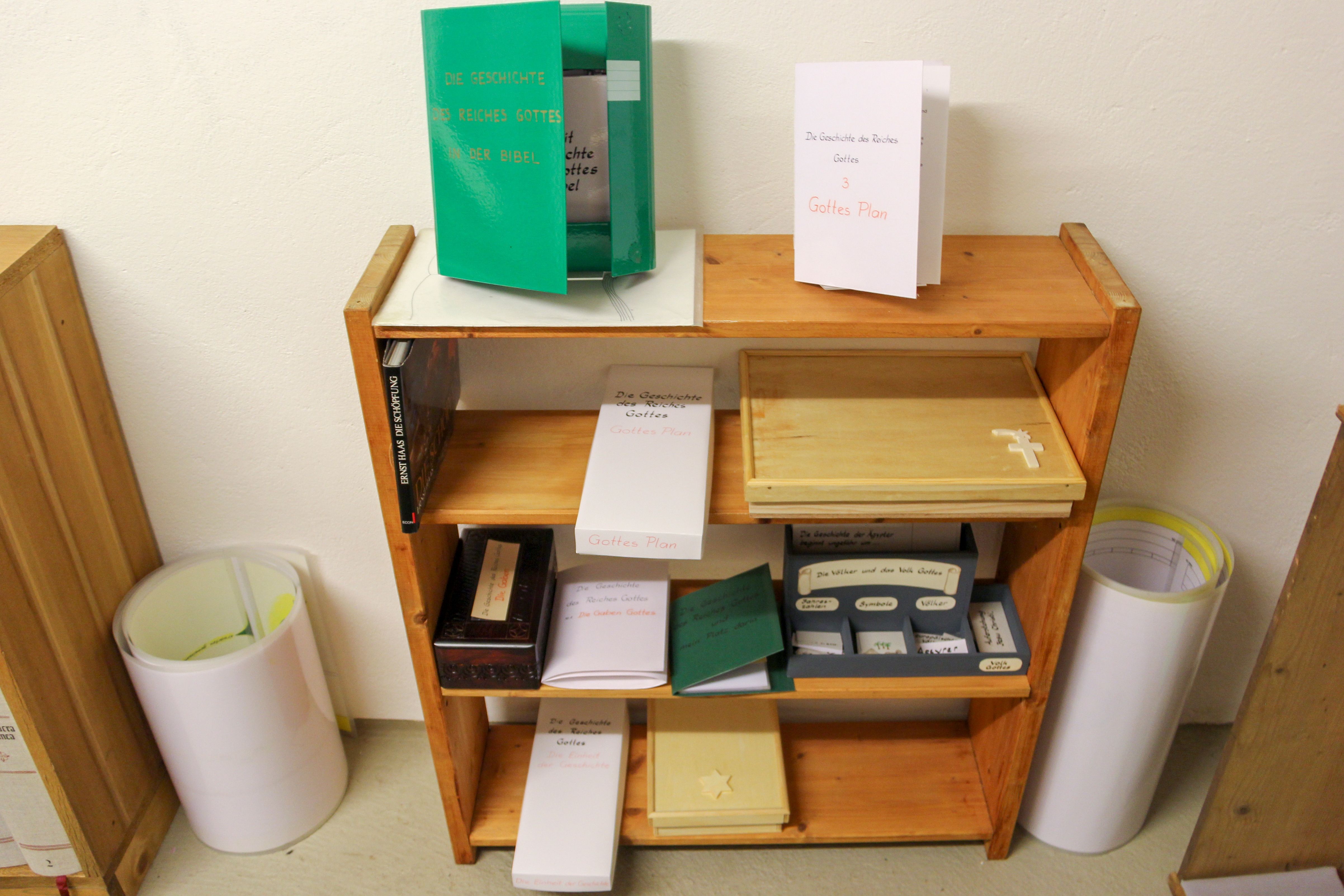 Offenes Regal mit Heften, Holzkisten, Legematerial und aufgerollten Bändern zur Geschichte des Reiches Gottes