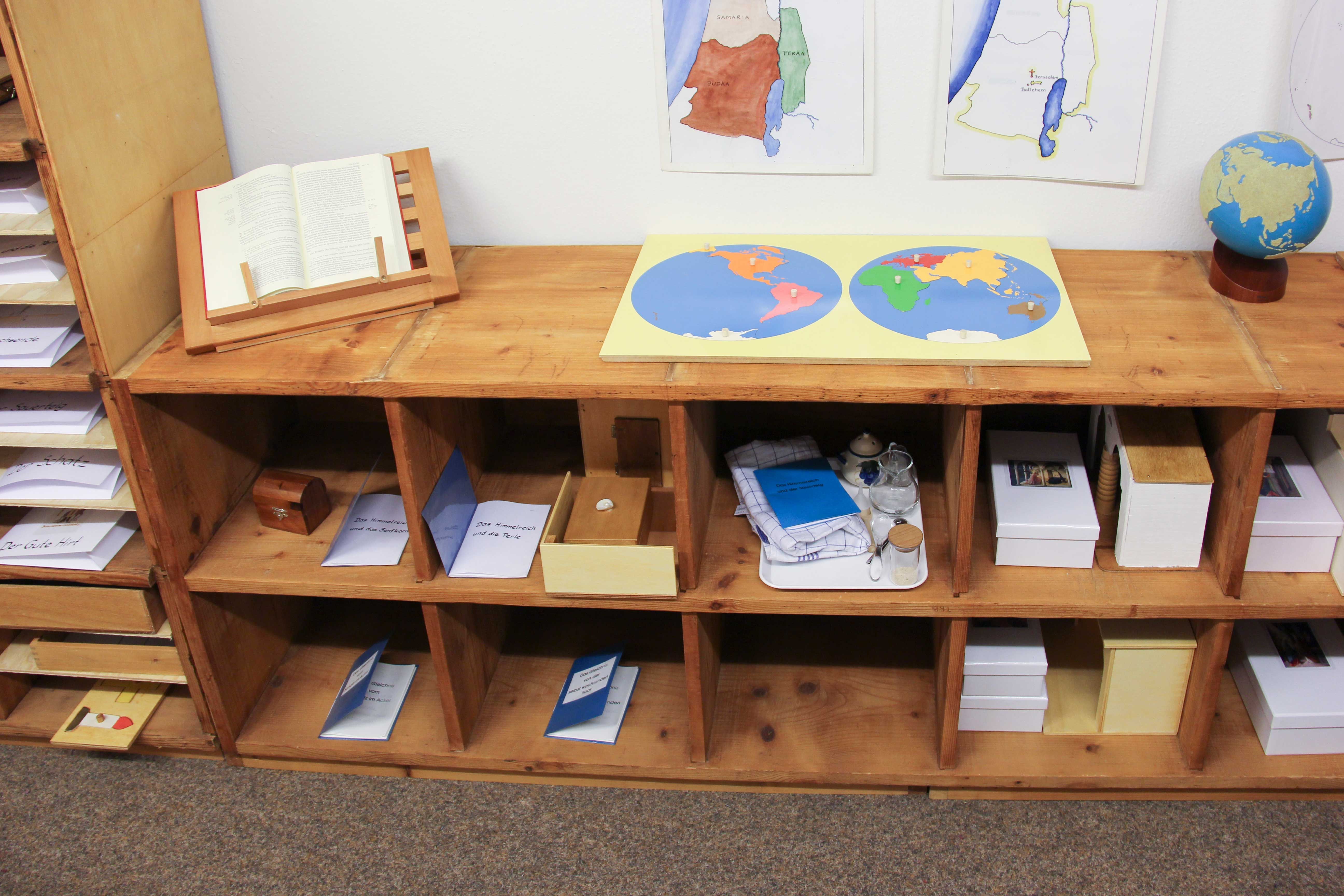 Offenes Regal mit Materialien über das Reich Gottes und Bibelstellen zur Kindheit Jesu, oben ein Sandpapierglobus und ein Puzzle der Erdteile