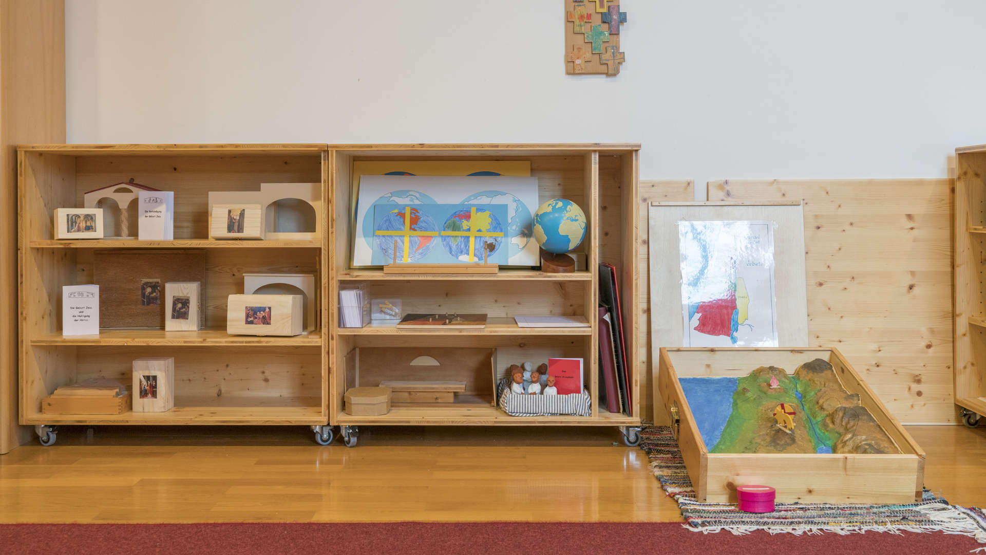 Offene, rollbare Regale mit kleinen Büchern mit Bibeltexten, dazu Figuren und kleinen Häusern, das Relief Israels und Landkarten