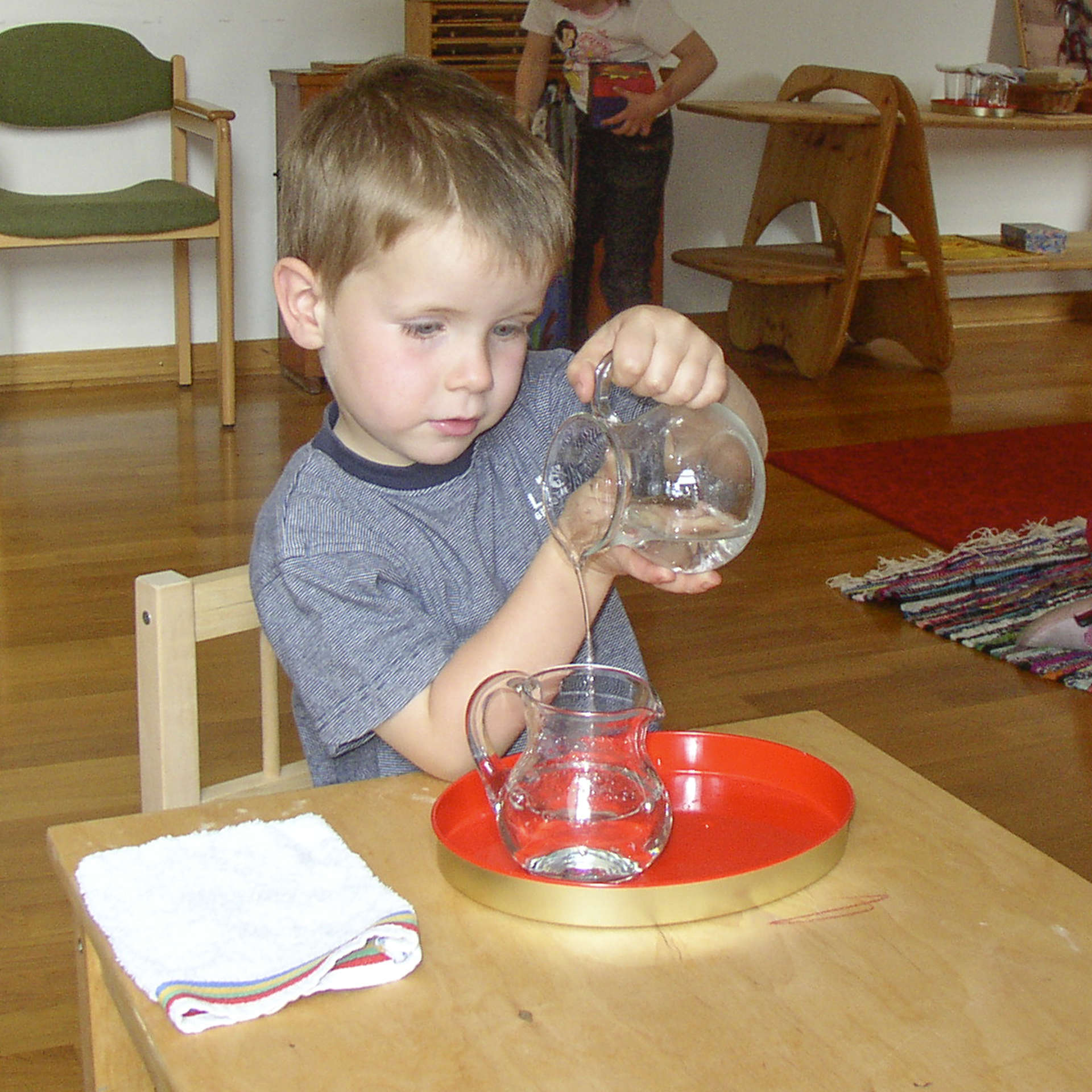 Kleiner Junge gießt konzentriert Wasser aus einem Kännchen in ein anderes Kännchen