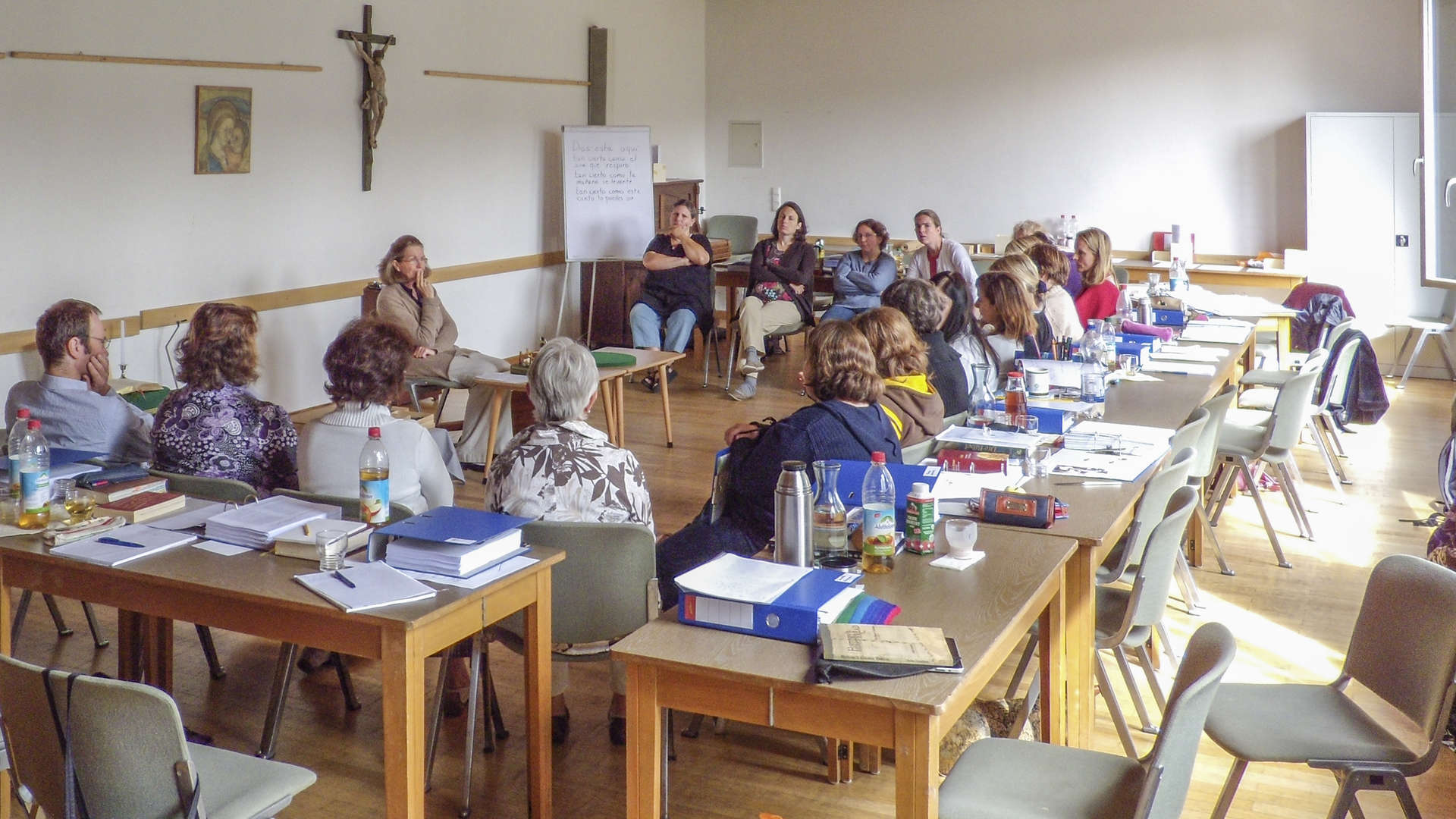 Pfarrsaal mit einem Tischkreis, innerhalb ein Stuhlkreis, die Kursteilnehmer hören bei der Präsentation eines Themas zu.